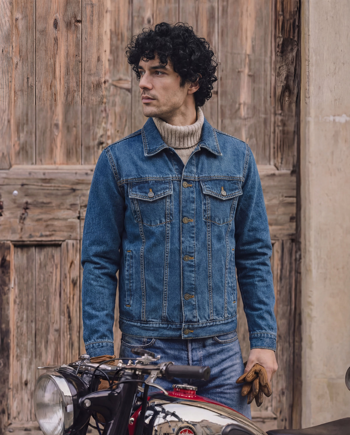 Man wearing a blue denim jacket standing next to a motorcycle with a wooden wall background