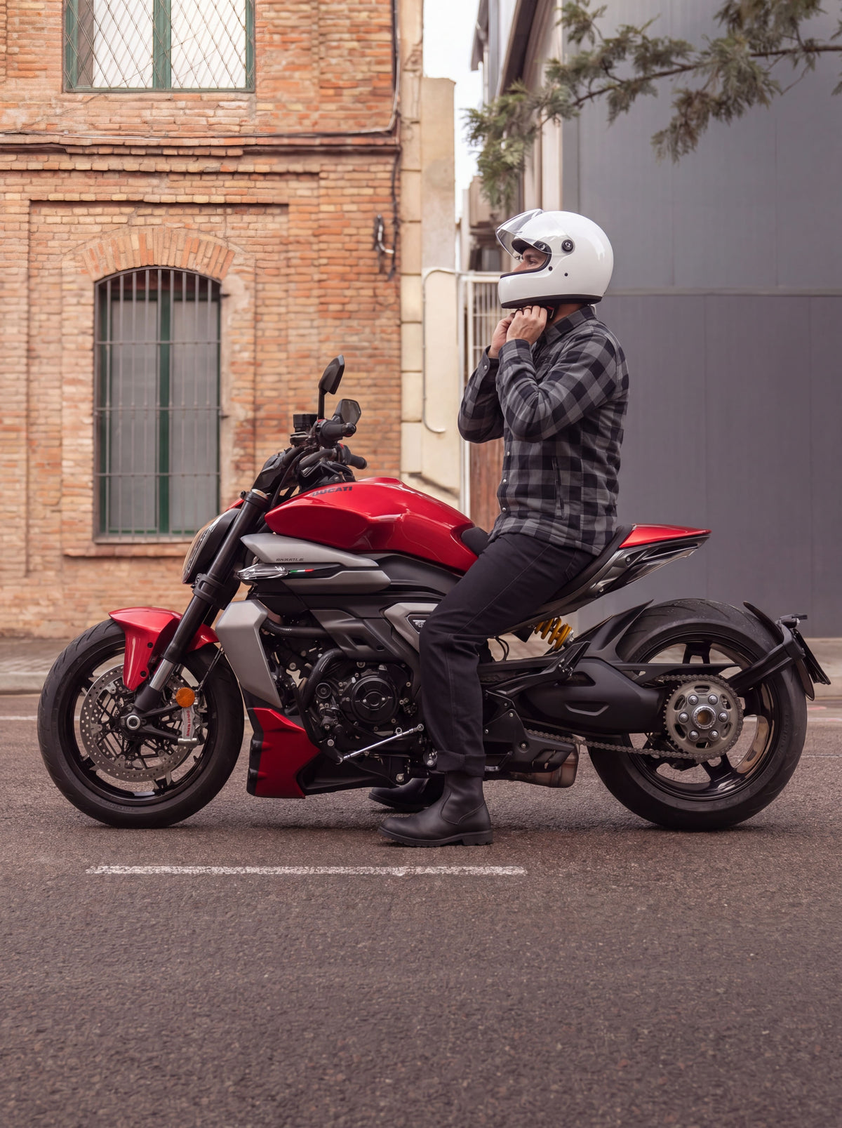 Rider in a grey and black motorcycle flannel shirt fastening a white helmet while seated on a red motorcycle.